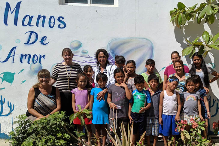 A group of adults and children pose for a photo in front of a white wall with the words Manos de Amor and painted ocean-themed decorations.