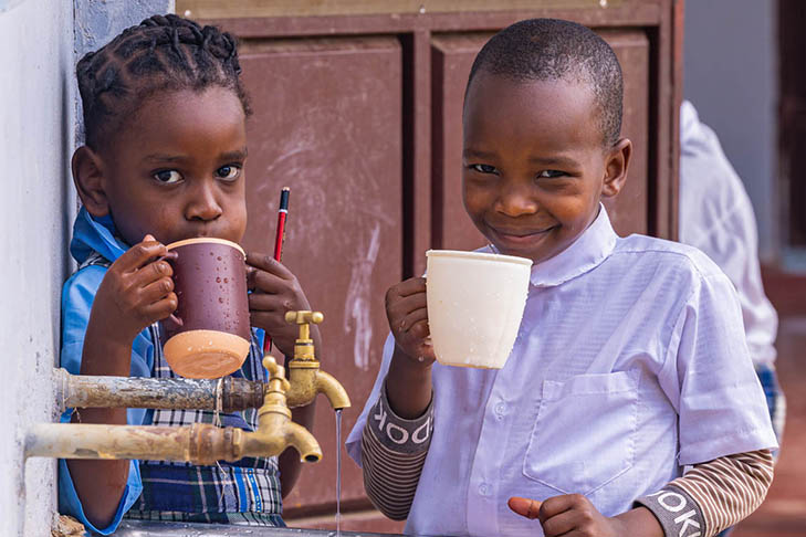 Two children standing near a water tap, holding and drinking from mugs, with one child wearing a school uniform and smiling at the camera.