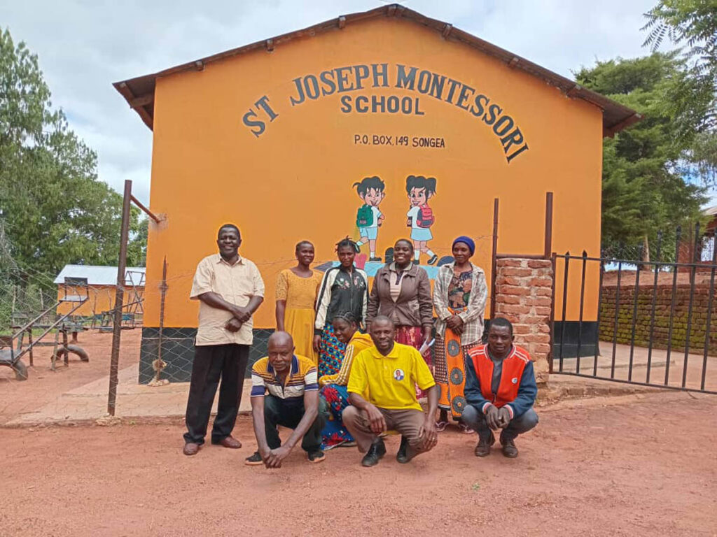 A group of nine adults pose for a photo outside a building labeled St Joseph Montessori School with cartoon children painted on the wall.