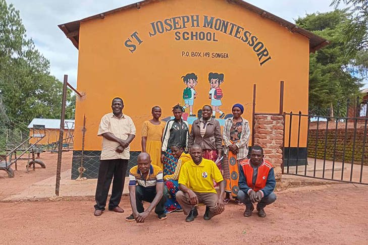 A group of nine adults pose in front of the yellow St Joseph Montessori School building with painted cartoon children and school name on the wall.