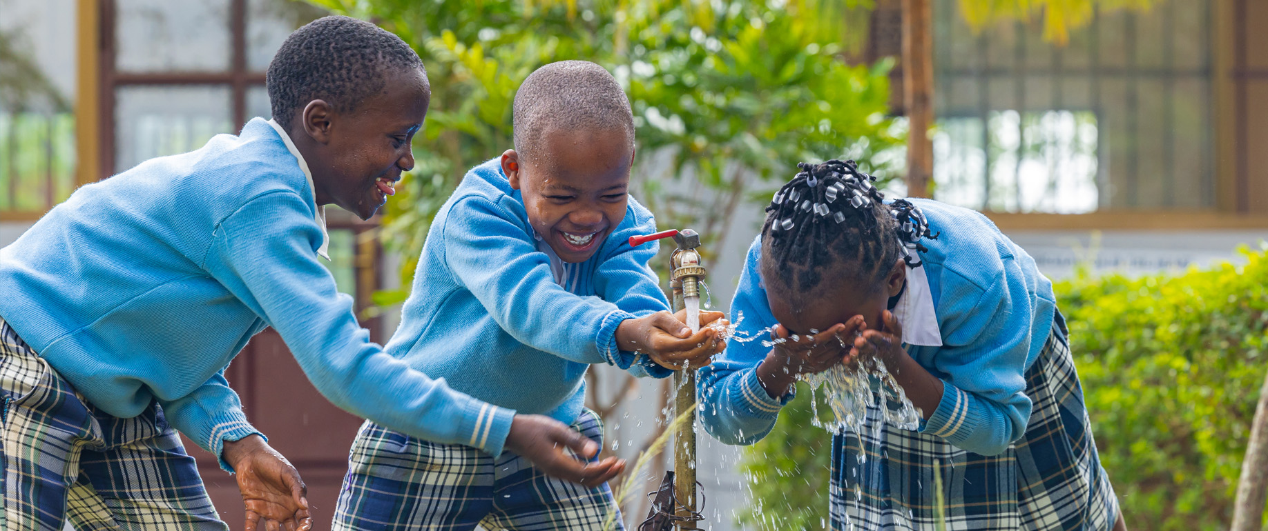 Three children in blue uniforms laugh and wash at an outdoor water tap during daytime, with greenery and a building in the background.
