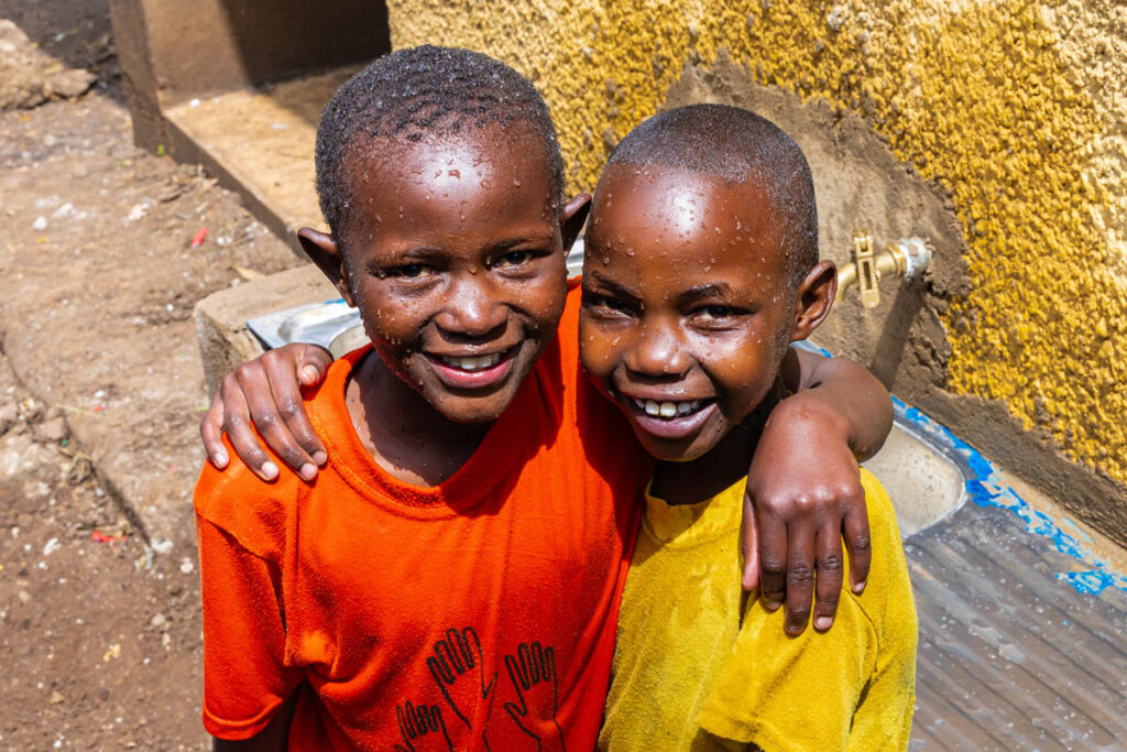 Two smiling children with wet faces and shirts stand arm-in-arm outdoors near a yellow wall and a water tap.