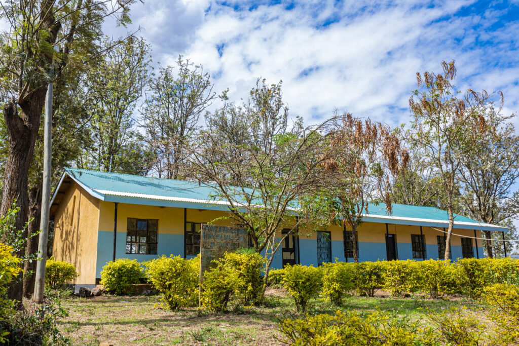 A single-story building with a blue-green roof and yellow and blue walls, surrounded by bushes and trees under a partly cloudy sky.