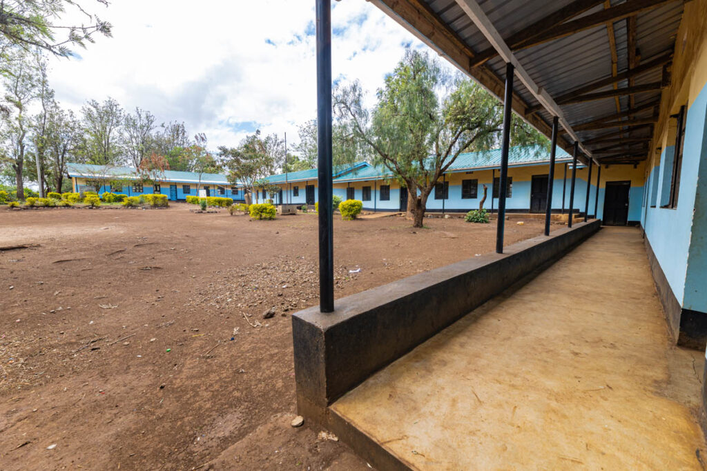 Outdoor view of a school courtyard with blue-painted buildings, a covered walkway, and scattered trees and shrubs.