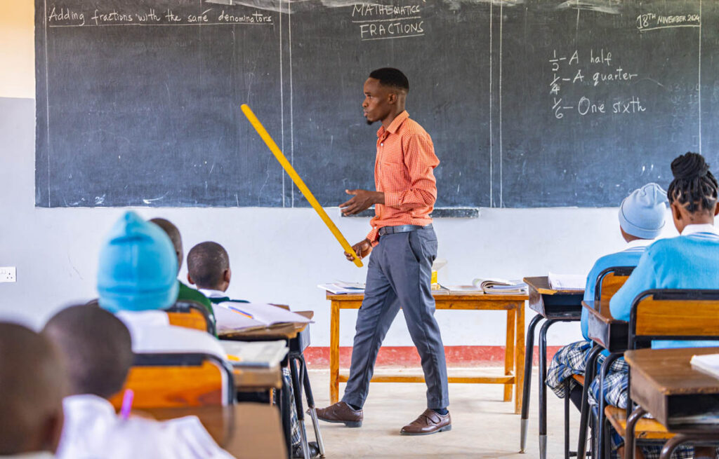 A teacher stands at the front of a classroom, pointing to a chalkboard with a yellow stick while students listen. The board displays math fractions and explanations.