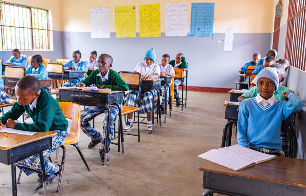 Students in uniform sit at their desks in a classroom, writing in notebooks. Educational posters are displayed on the walls behind them.