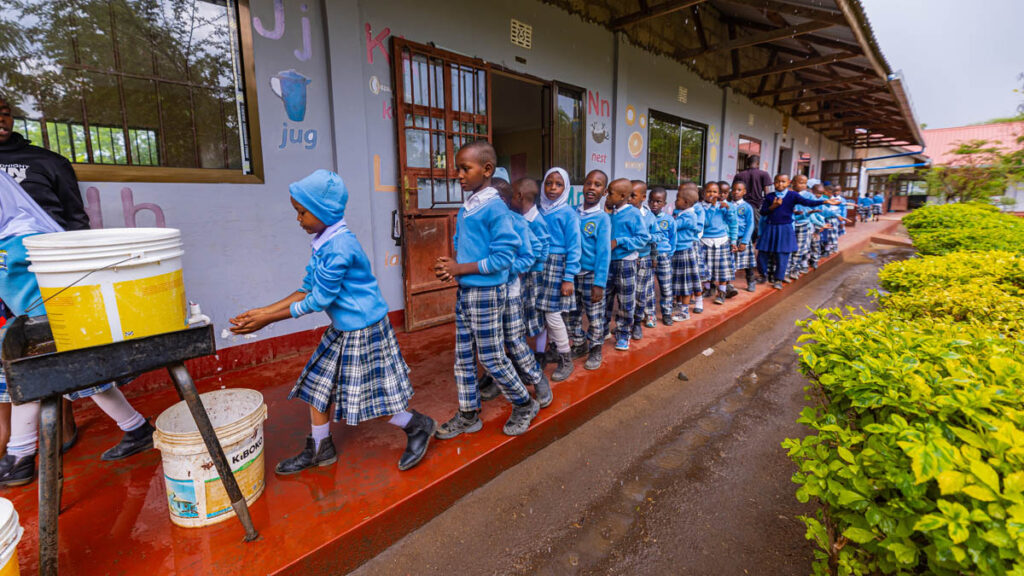 A group of schoolchildren in uniform stand in a line outside a classroom building, waiting to wash their hands at a handwashing station.
