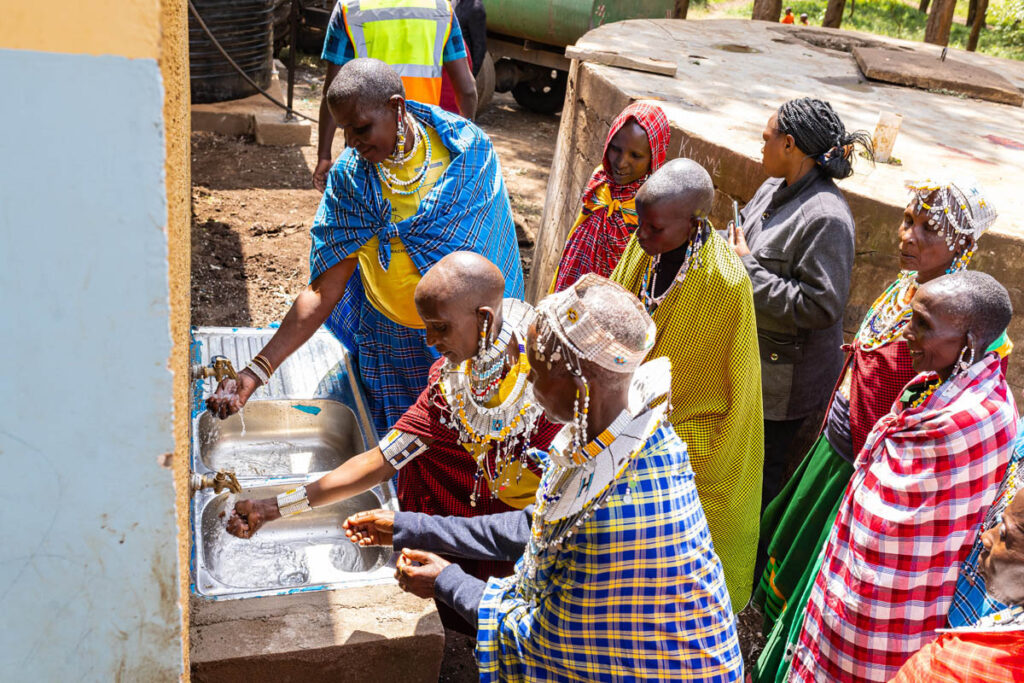 A group of people in colorful traditional clothing use a communal outdoor sink to wash their hands.