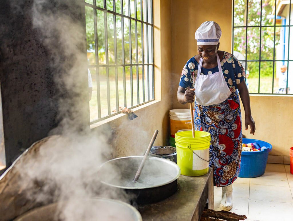 A woman wearing an apron and headscarf stirs a large pot with steam rising, inside a kitchen with large windows and buckets on the floor.