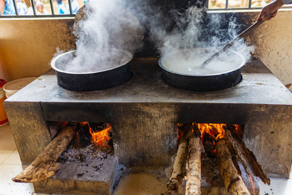 Two large pots on a wood-fired stove with burning logs and steam rising, while a person stirs one of the pots.