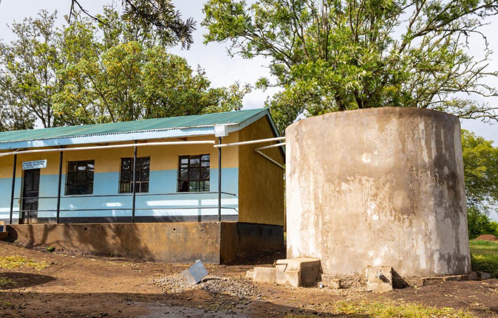 A concrete water tank stands next to a single-story building with a tin roof and blue walls, surrounded by trees and dirt ground.