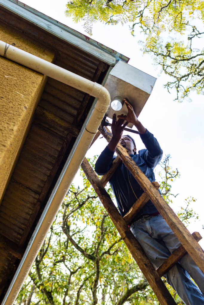 A person stands on a wooden ladder, installing or repairing a pipe connected to the gutter of a building, with trees visible in the background.