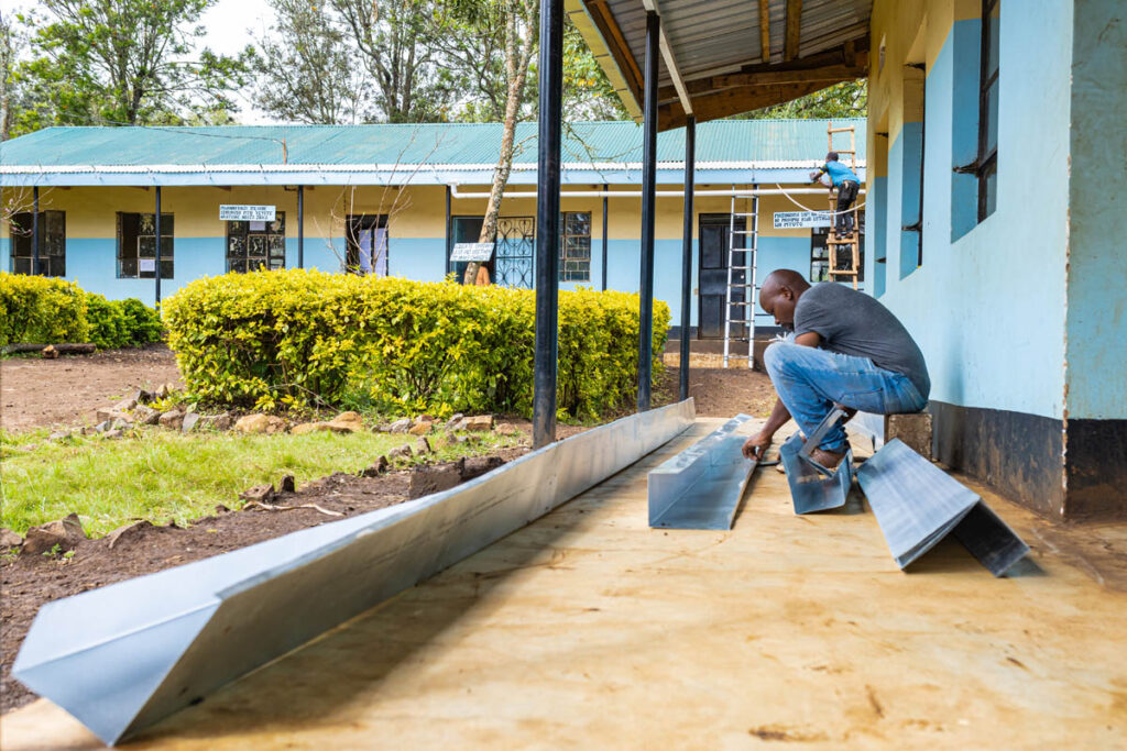 A man installs metal gutters on the ground outside a blue building, with more gutters and greenery visible in the background.