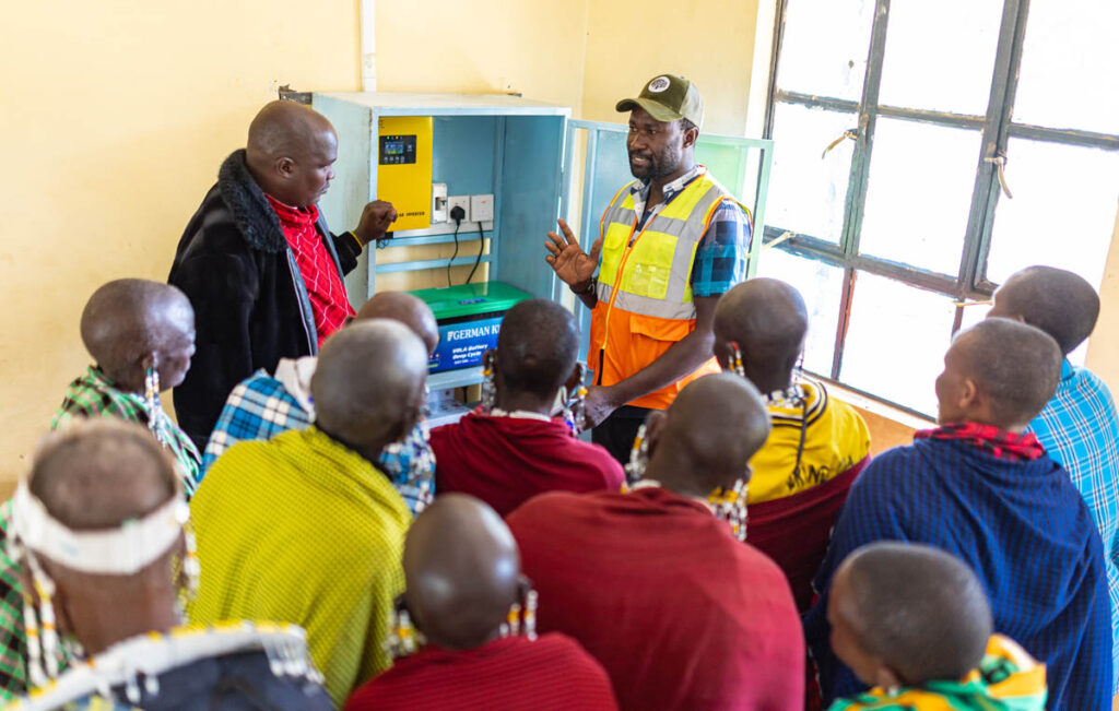 A group of people observe two men demonstrating the use of an electrical device inside a room with a window.