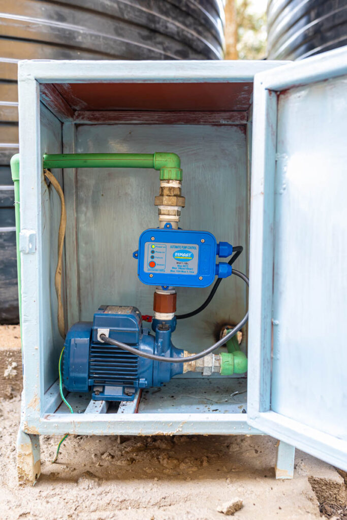 A water pump with attached control unit is installed inside a light blue metal cabinet with an open door; green pipes connect to the pump.