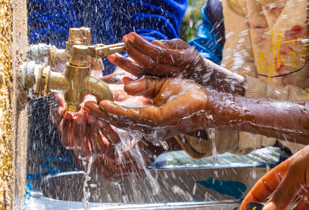 Multiple hands are washing under running water from a brass outdoor tap, with water splashing around them.