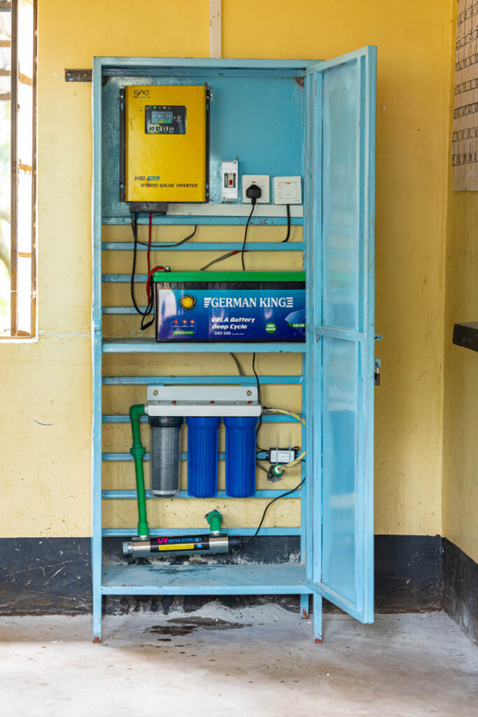 A blue metal cabinet contains a solar inverter, battery, water filtration system, and related electrical components mounted on a yellow wall.