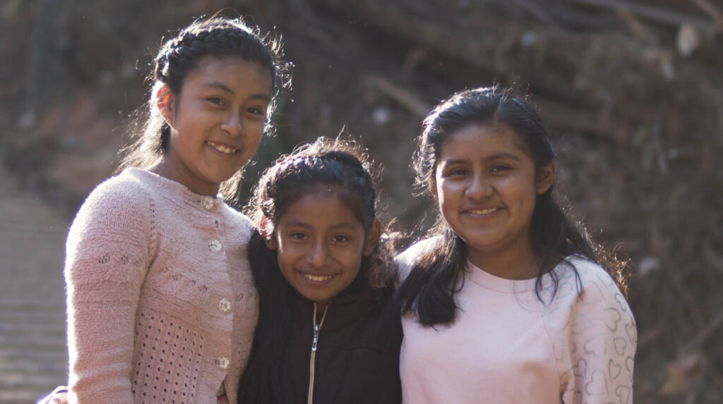 Three girls stand close together outdoors, smiling at the camera. They are wearing sweaters and are lit by natural sunlight.