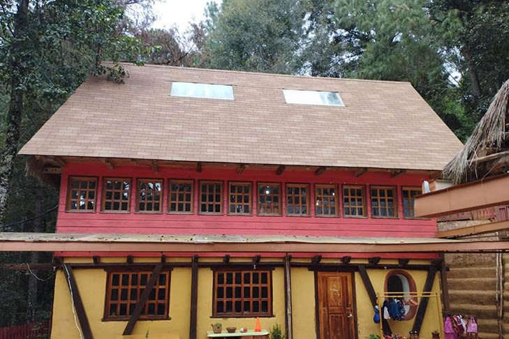 Two-story house with a yellow lower level and red upper level, wooden framed windows, steep roof, and surrounded by trees.