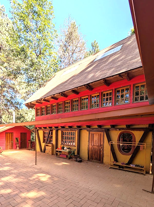A two-story building with red and yellow trim, wooden doors, and multiple windows, surrounded by trees and a brick-paved courtyard on a sunny day.