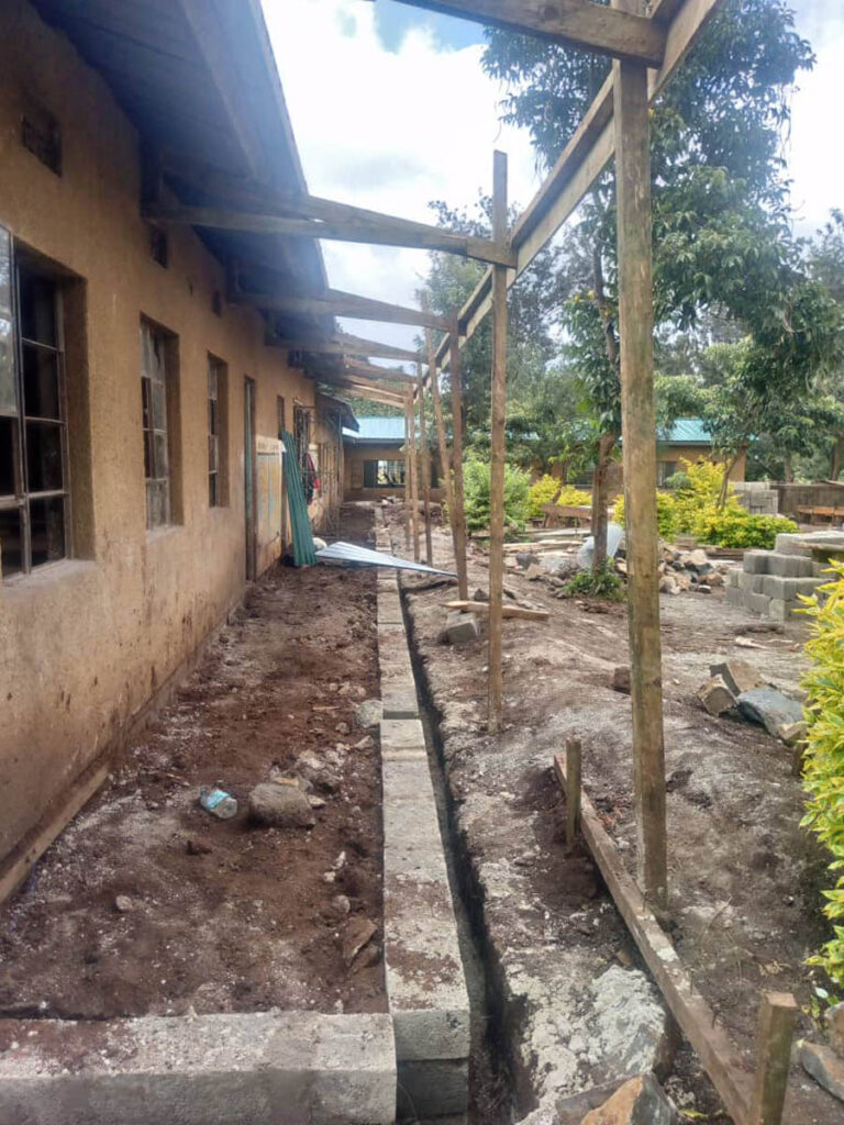 A construction site beside a building with a dug trench, wooden supports, scattered rocks, and some green trees and shrubs nearby.