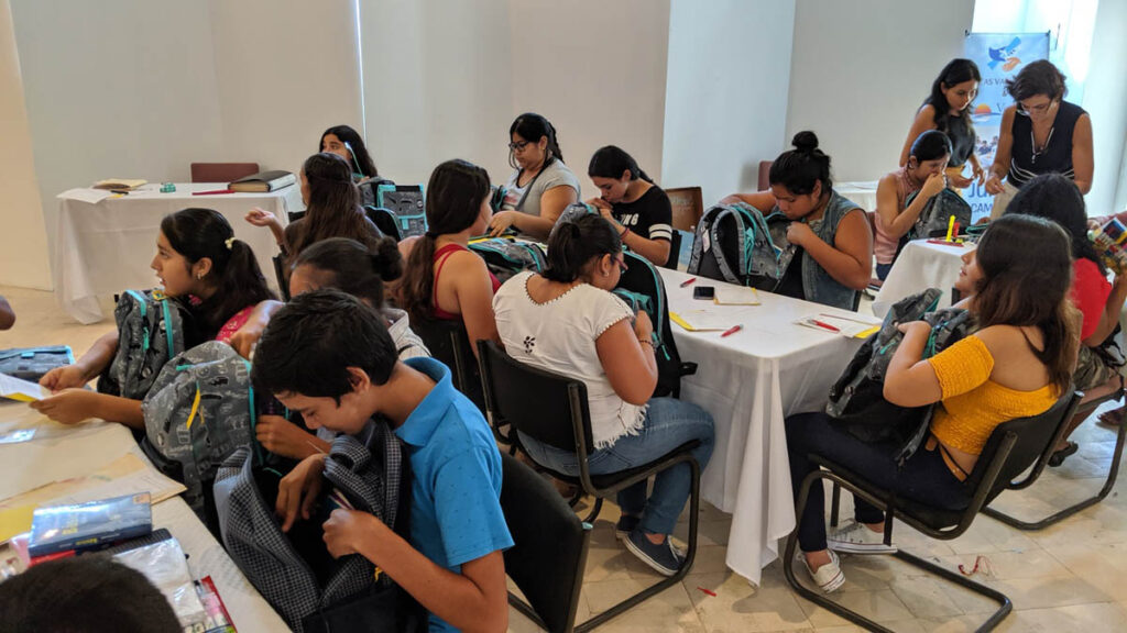 A group of students sitting at tables in a classroom, sorting through school supplies and backpacks.
