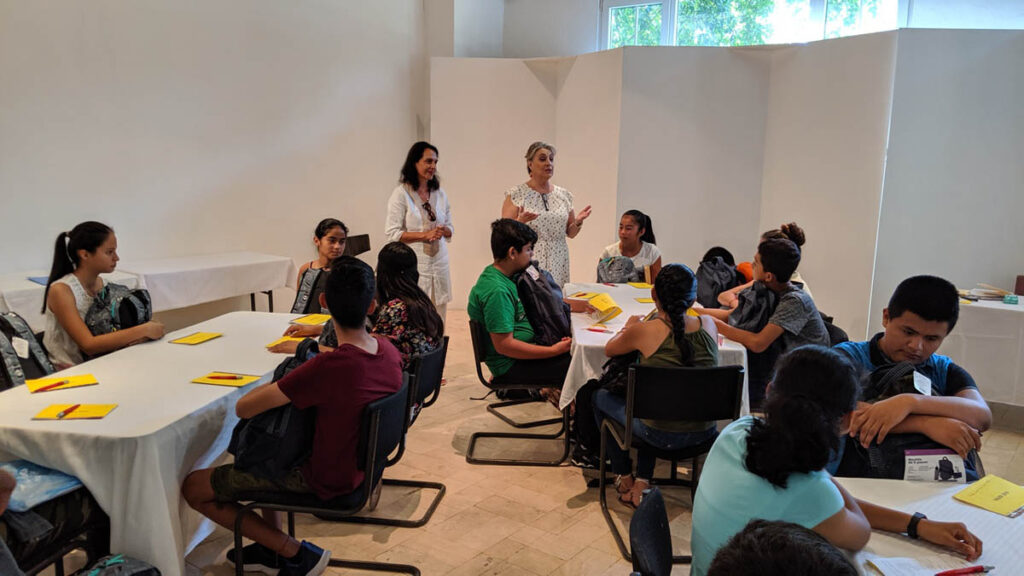 Two women stand and speak to a group of students seated at round tables in a classroom, with yellow papers in front of each student.