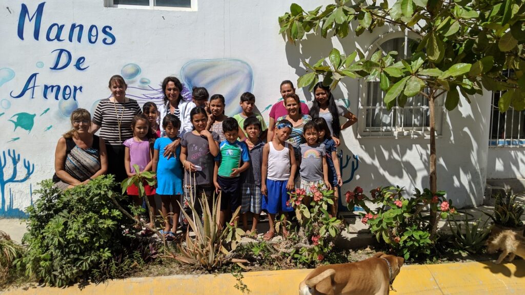 A group of adults and children stand together in front of a white building with Manos de Amor painted on the wall, surrounded by plants, with a dog in the foreground.