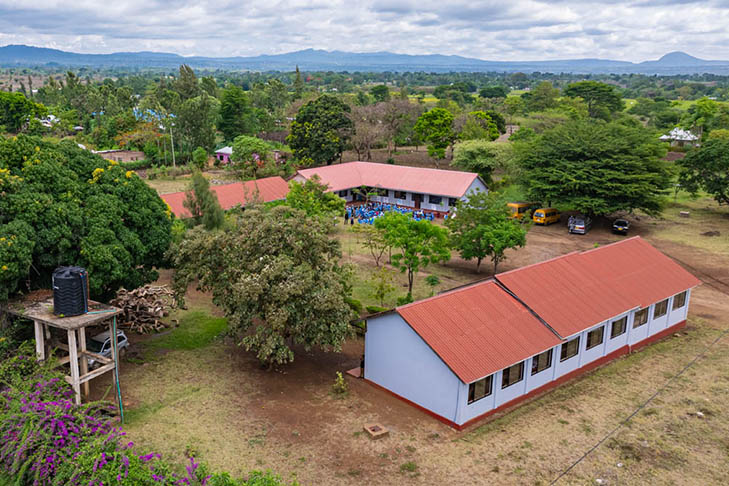Aerial view of a rural school with red-roofed buildings, trees, open grounds, and distant hills under a cloudy sky.