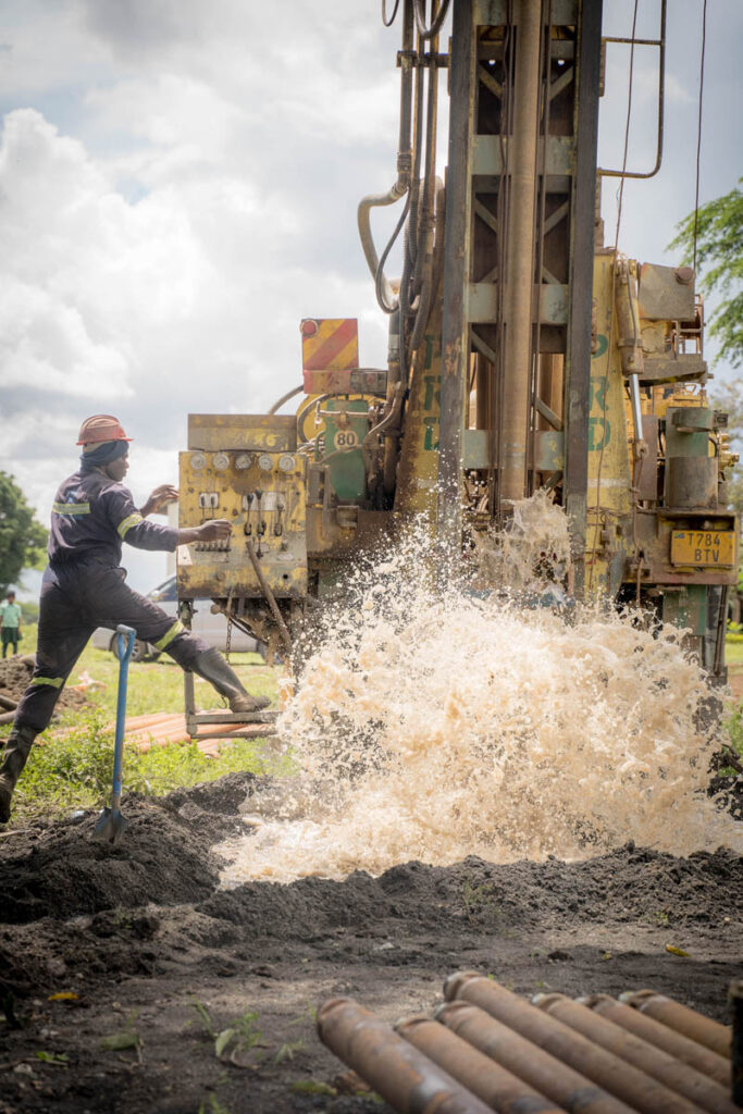 A worker in safety gear operates a large drilling machine as water and mud spray from the ground during a borehole drilling operation.