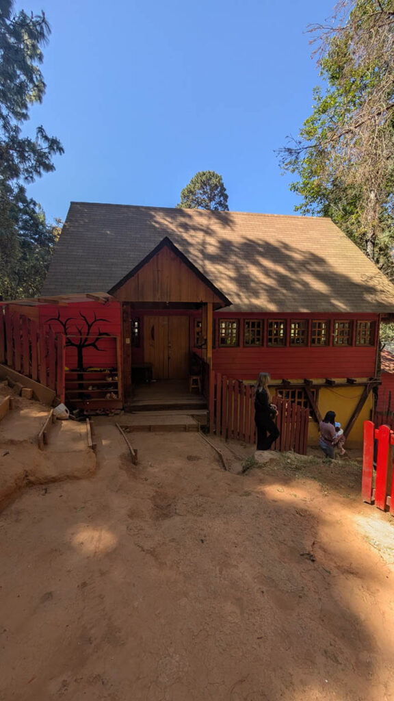 A wooden building with red accents sits among trees on a dirt path; two people are near the entrance and steps lead up to the house.