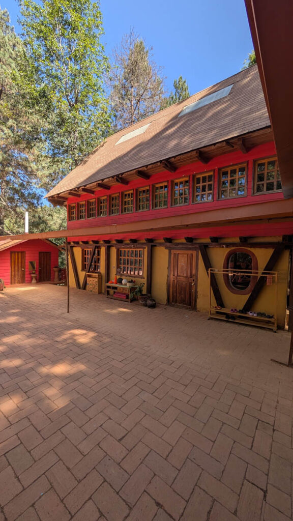 A two-story building with red-framed windows and a sloped roof, surrounded by trees and a paved courtyard on a sunny day.