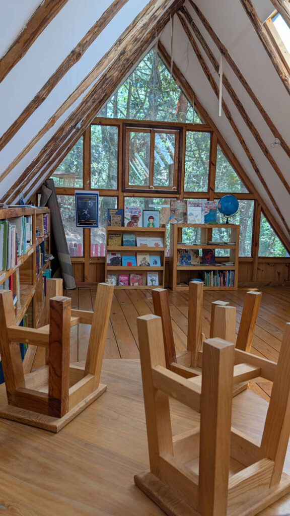 A cozy, wooden, attic-style library with bookshelves, children’s books, a globe, and chairs stacked upside down on a round table. Large triangular windows show green trees outside.