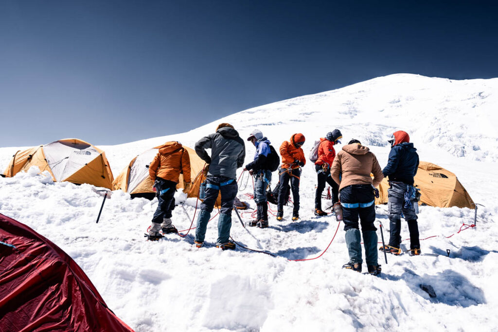 A group of climbers wearing winter gear stands on snow near yellow tents at a high-altitude mountain campsite under a clear blue sky.