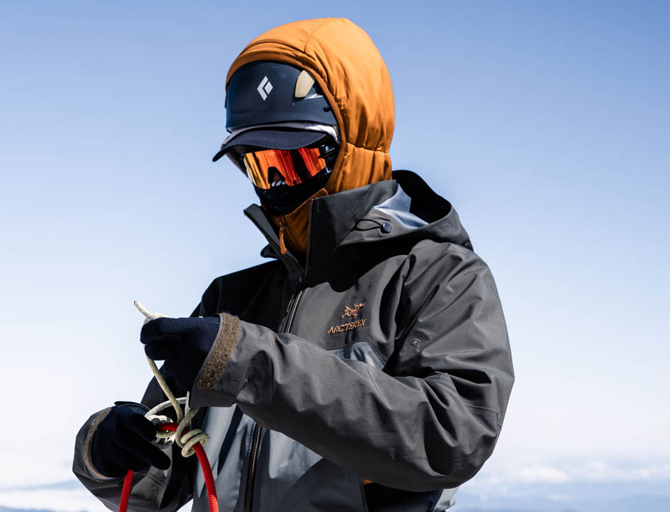 A person in winter outdoor gear ties a knot in a rope, with a snowy mountain landscape in the background.