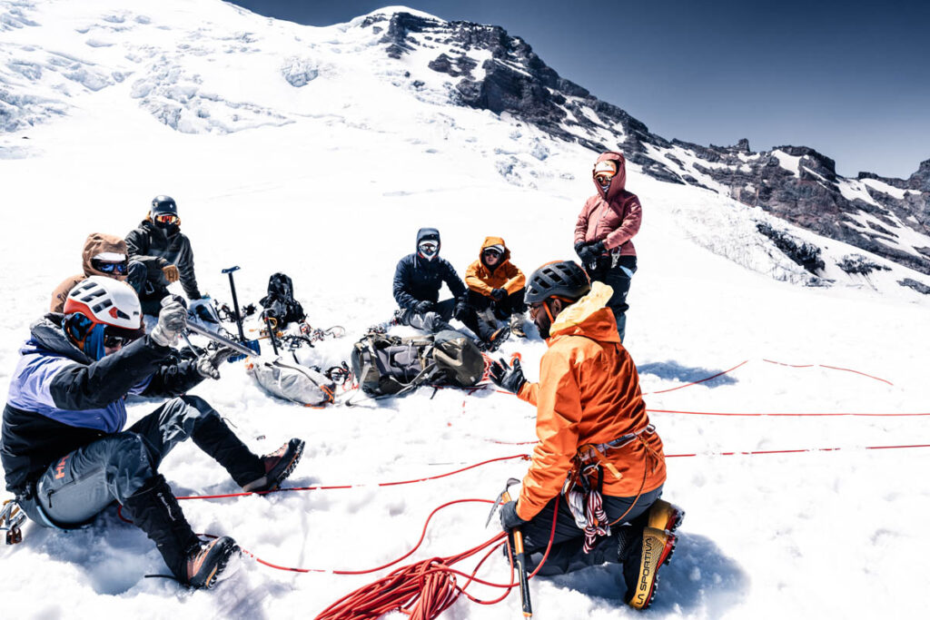 A group of mountaineers in winter gear sit on snow with climbing equipment and ropes, resting on a snowy mountain slope under clear skies.