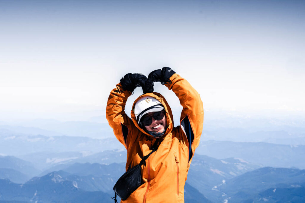 A person in an orange jacket and sunglasses stands on a mountain summit, smiling and making a heart shape with their arms above their head. Mountain landscape is visible in the background.