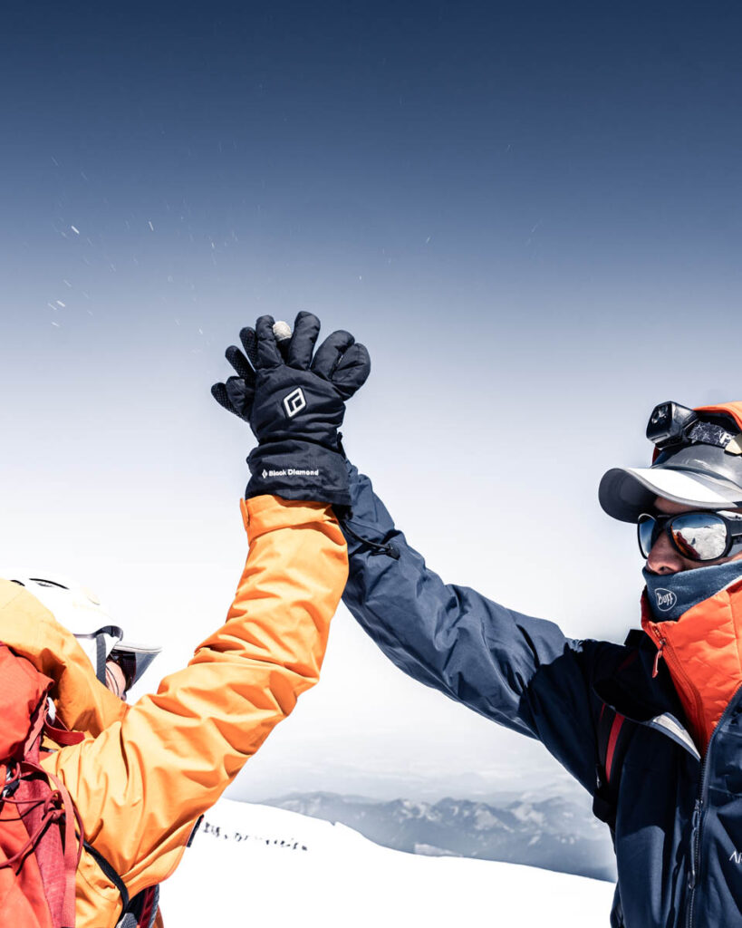 Two climbers wearing protective gear and gloves high-five each other on a snowy mountain summit under a clear sky.