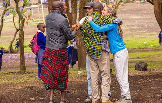 Four adults, including two in traditional Maasai clothing, greet each other warmly in an outdoor setting with children in school uniforms in the background.