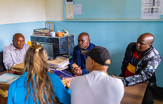 Five people are seated around a desk in an office, engaged in a discussion. There are folders, papers, and office supplies on the desk.