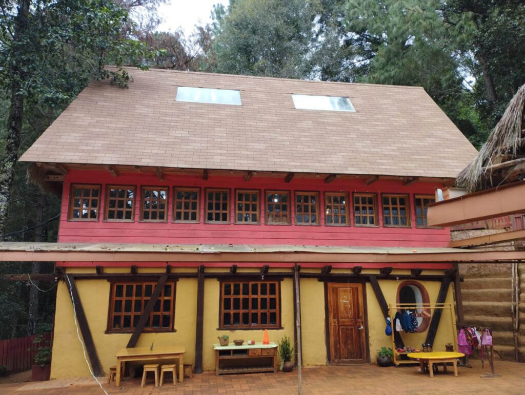 A two-story building with a yellow lower level and a red upper level, wooden windows, a sloped roof with skylights, and benches in front, surrounded by trees.