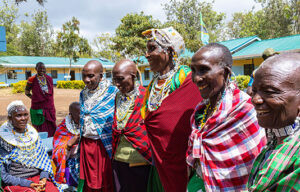 A group of Maasai women in traditional clothing and beadwork stand and sit together outdoors, smiling, with buildings and trees in the background.