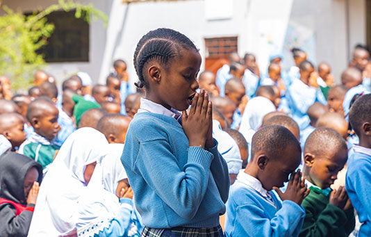 A group of schoolchildren in uniform stand outdoors with their hands clasped in prayer.