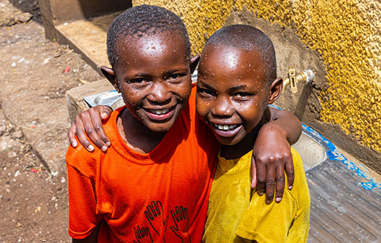 Two smiling children with wet faces, one in an orange shirt and the other in yellow, stand together with arms around each other near a water tap against a yellow wall.