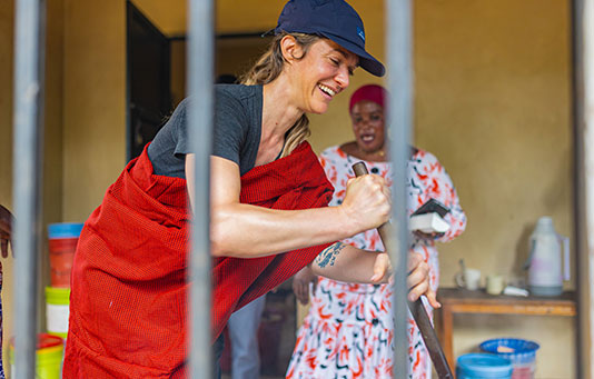 A woman smiling and stirring food in a large container indoors, with another woman in patterned clothing standing in the background. Vertical metal bars are visible in the foreground.