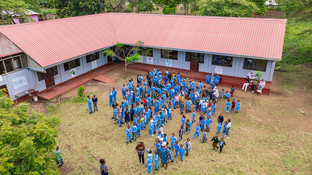 Aerial view of a group of children and adults in blue uniforms gathered outside a building with a red roof on a grassy area.