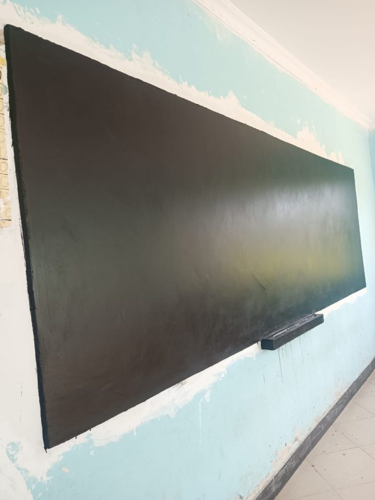 A large, freshly painted blackboard mounted on a light blue wall with paint marks around the edges; a narrow ledge runs along the bottom of the blackboard.