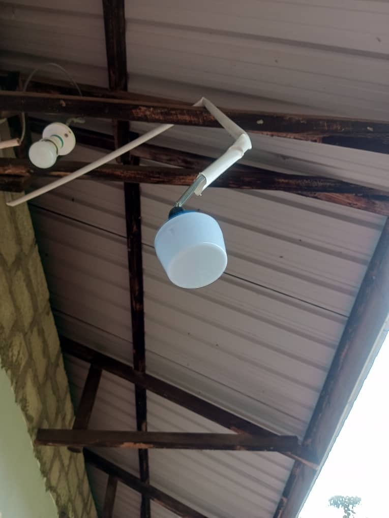 A white and blue security camera mounted on a metal beam under a corrugated metal roof, near an exposed electrical conduit and wall.