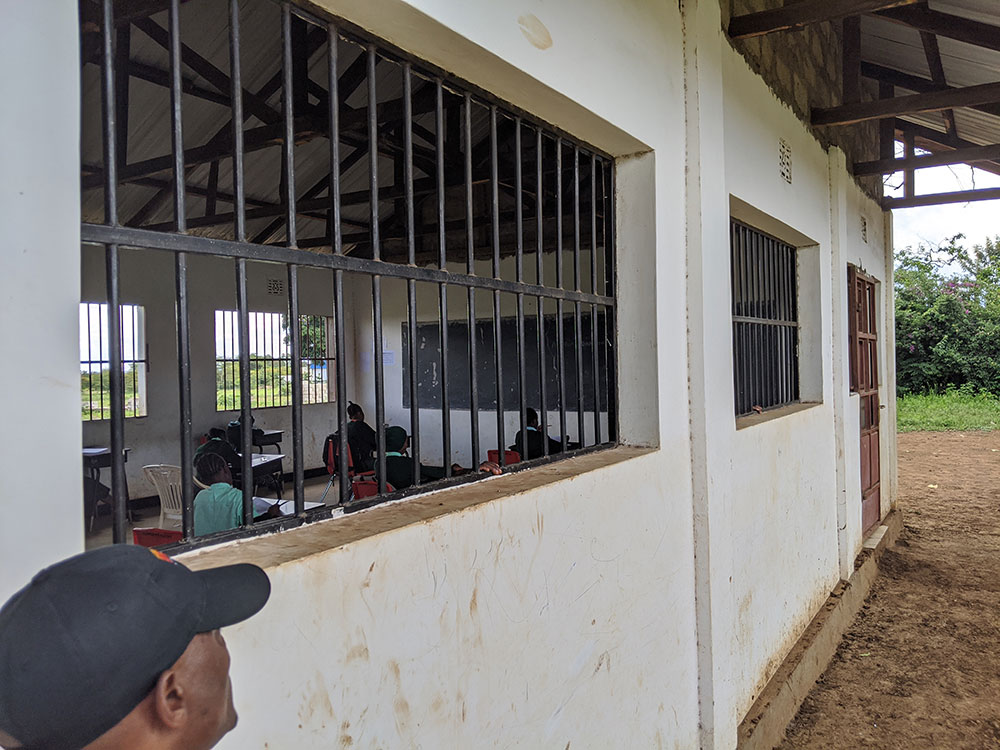 A man stands outside a classroom with barred windows, looking in at students seated at desks with a chalkboard at the front of the room.