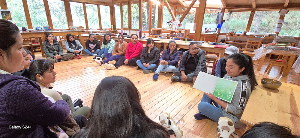 A group of people sits in a circle on a wooden floor inside a large room as one person reads aloud from a picture book.